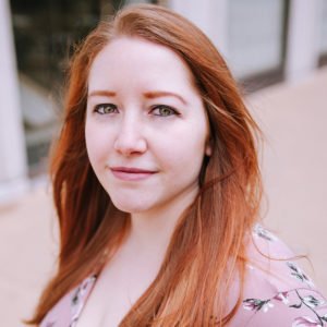 Tessa Watkins (they/them) present in this photo as a white person with long red hair, green eyes, and a pink floral blouse.