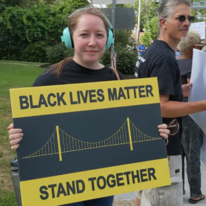 Tessa standing on the sidewalk with other protestors wearing ear defenders and holding a sign that reads, "Black Lives Matter | Stand Together" depicting a Pittsburgh bridge.